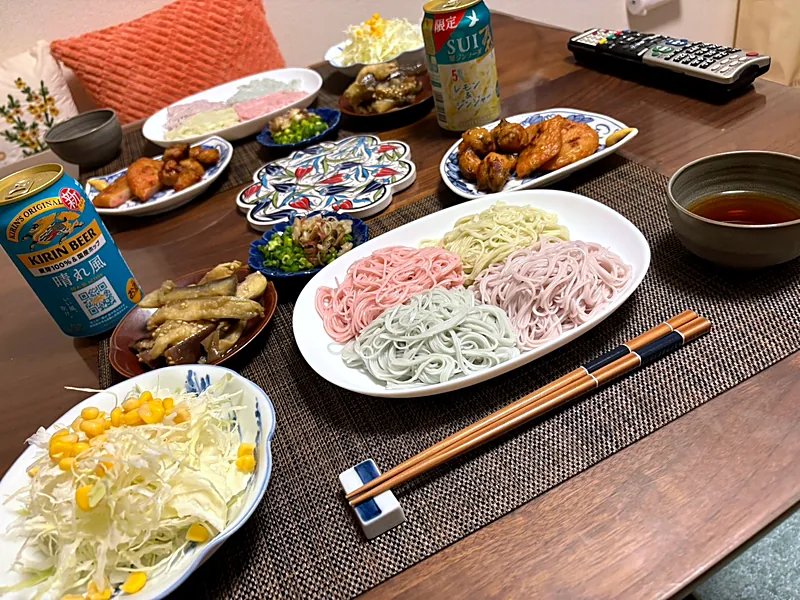 A platter of somen noodles in four colors with dipping sauce, plates of fried chicken, eggplant, cabbage salad with corn, and canned drinks