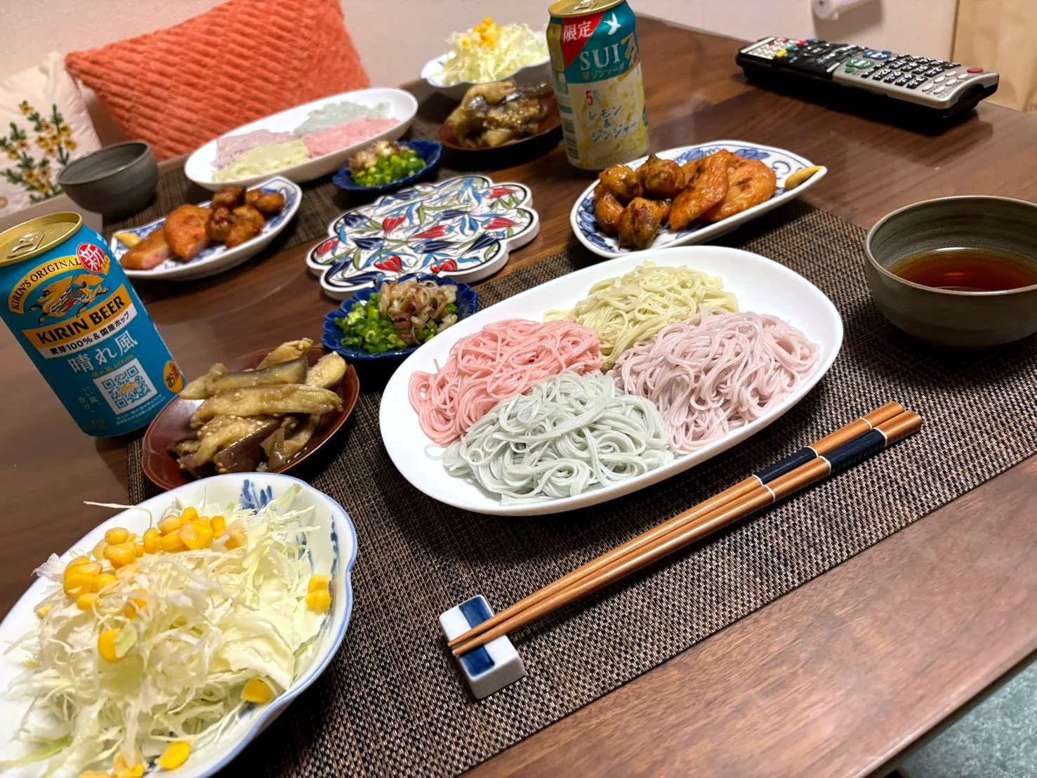 A platter of somen noodles in four colors with dipping sauce, plates of fried chicken, eggplant, cabbage salad with corn, and canned drinks