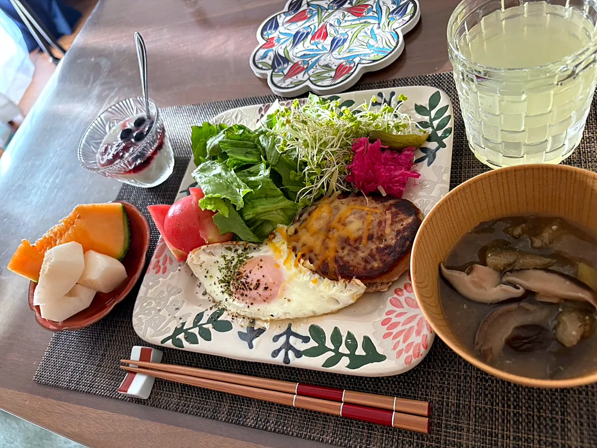 Hamburger steak with melted cheese and a fried egg beside lettuce and sprouts, with mushroom soup in a wooden bowl, melon and yogurt