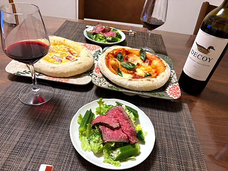 Two homemade pizzas, a salad topped with sliced roast beef, and glasses of red wine on a weekend table