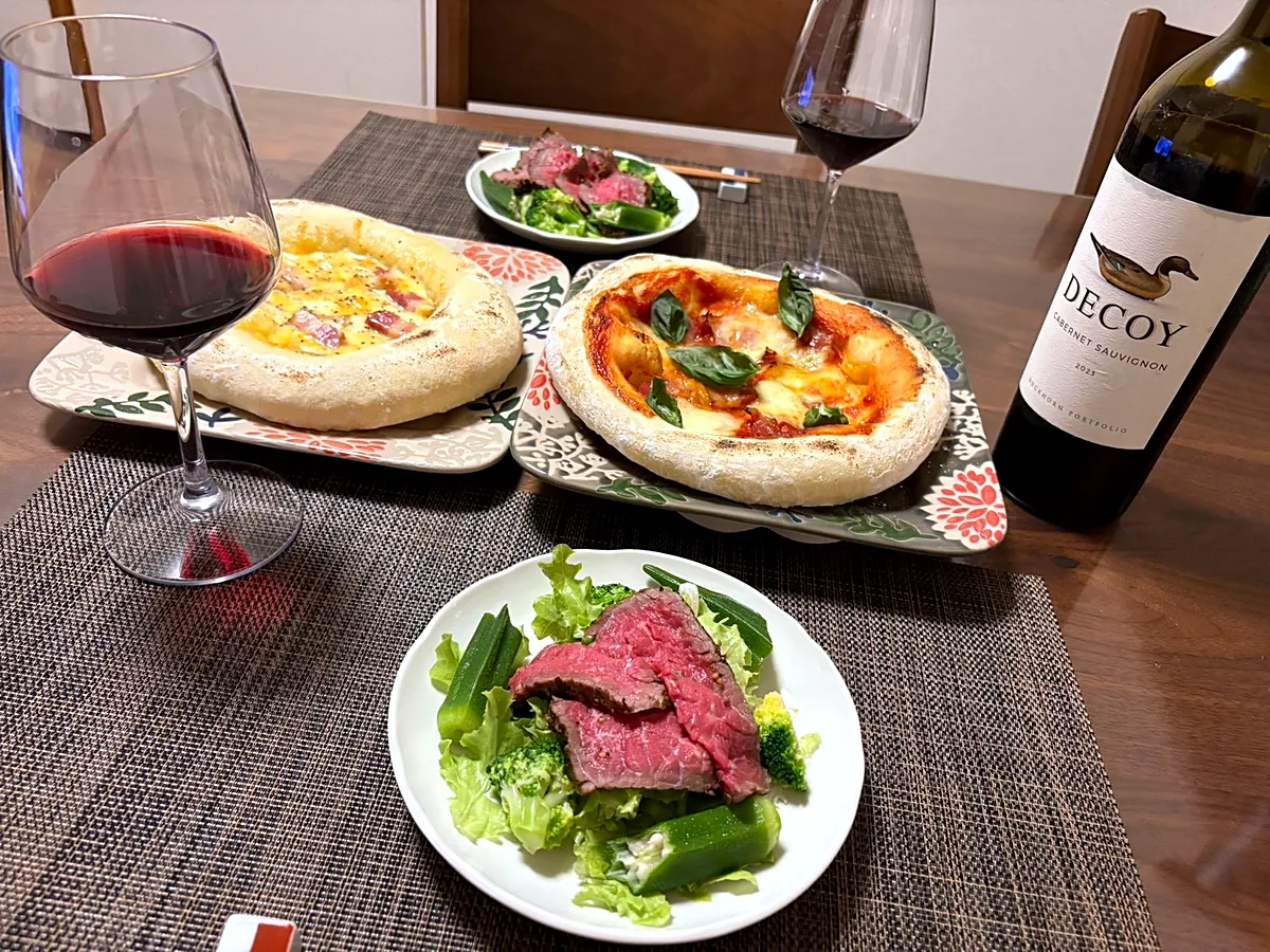 Two homemade pizzas, a salad topped with sliced roast beef, and glasses of red wine on a weekend table