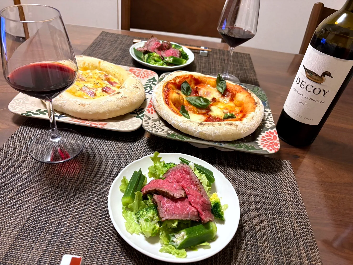 Two homemade pizzas, a salad topped with sliced roast beef, and glasses of red wine on a weekend table