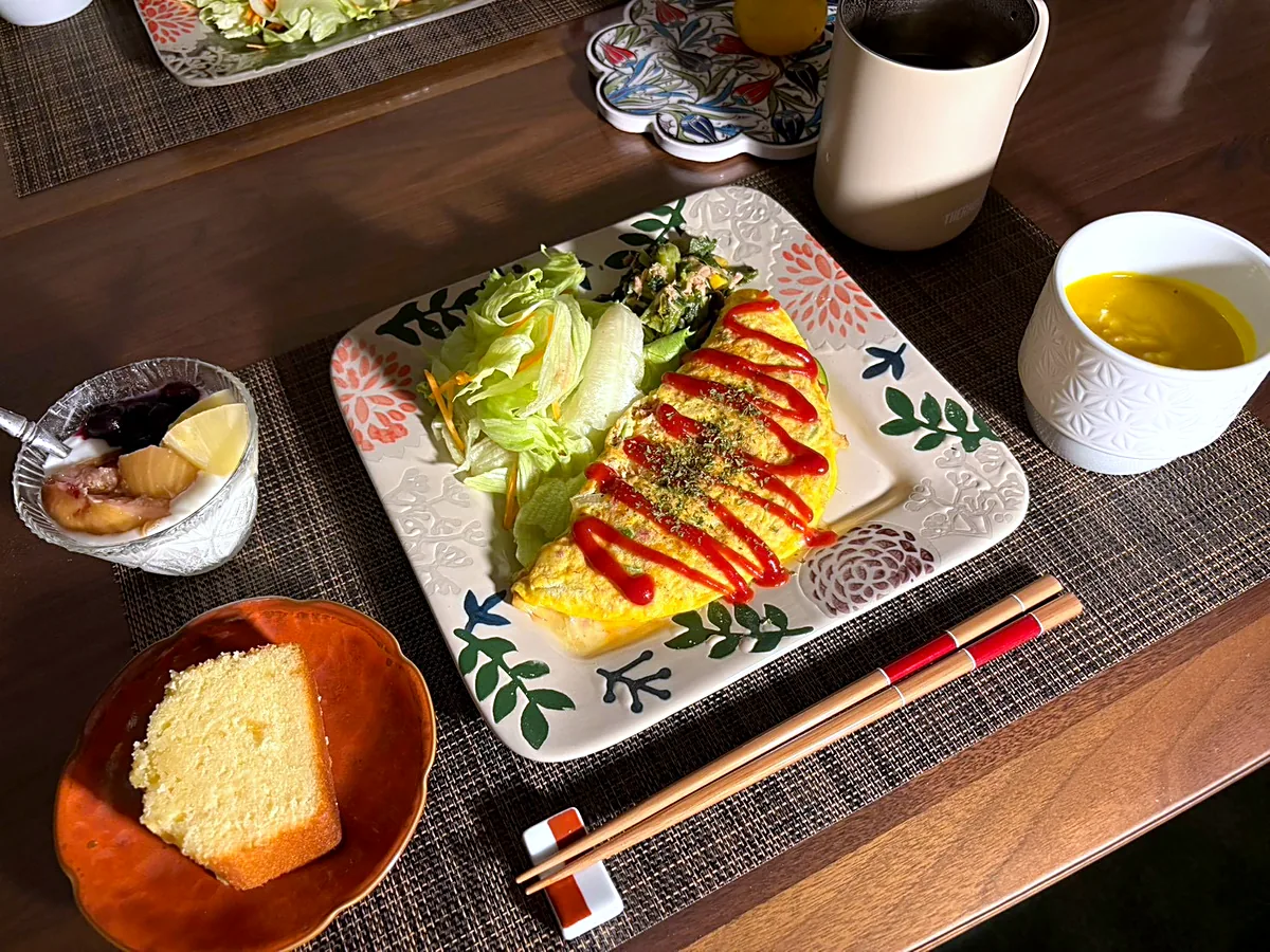 Omurice topped with ketchup beside a green salad, with a cup of pumpkin soup, a slice of pound cake and yogurt with fruit