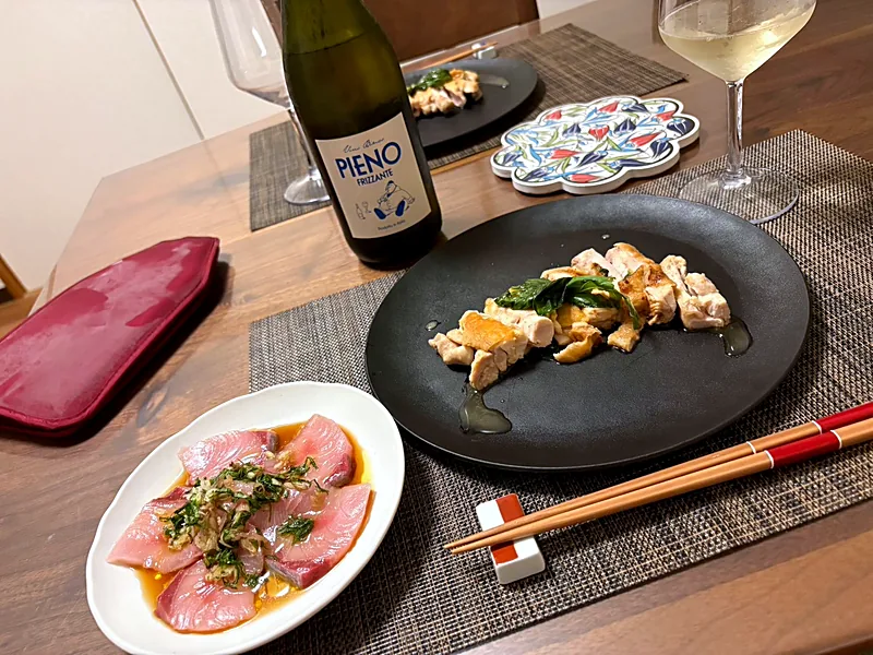 Slices of sashimi dressed with herbs on a white plate, sautéed chicken on a black plate, and white wine
