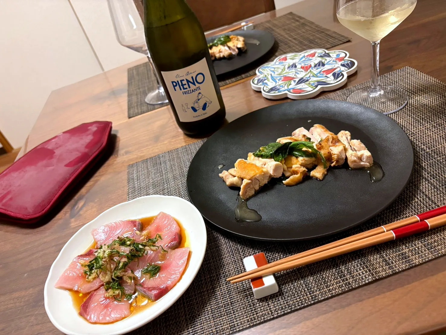 Slices of sashimi dressed with herbs on a white plate, sautéed chicken on a black plate, and white wine
