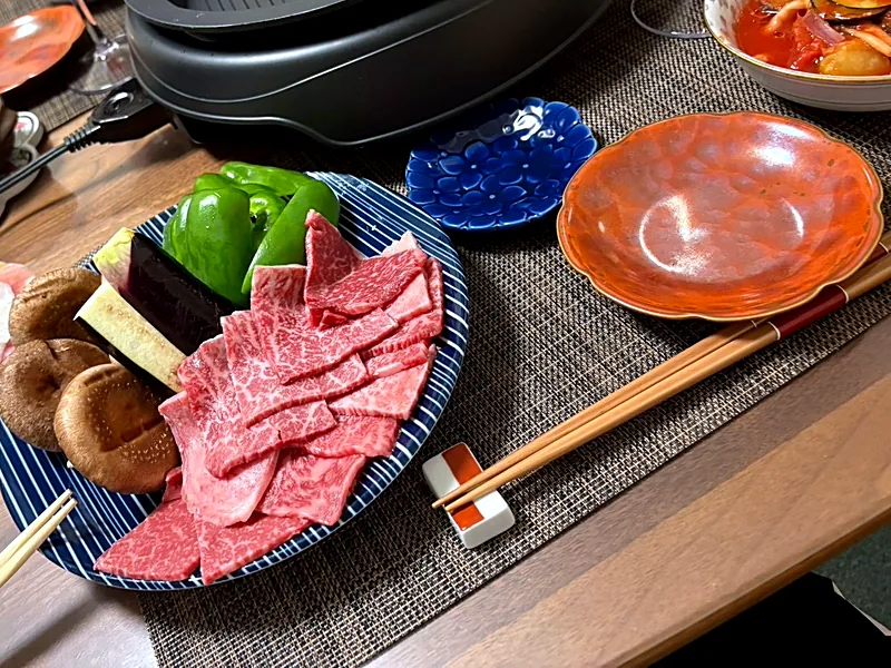 Raw beef and pork slices, shiitake mushrooms, green peppers and eggplant on plates beside a tabletop grill