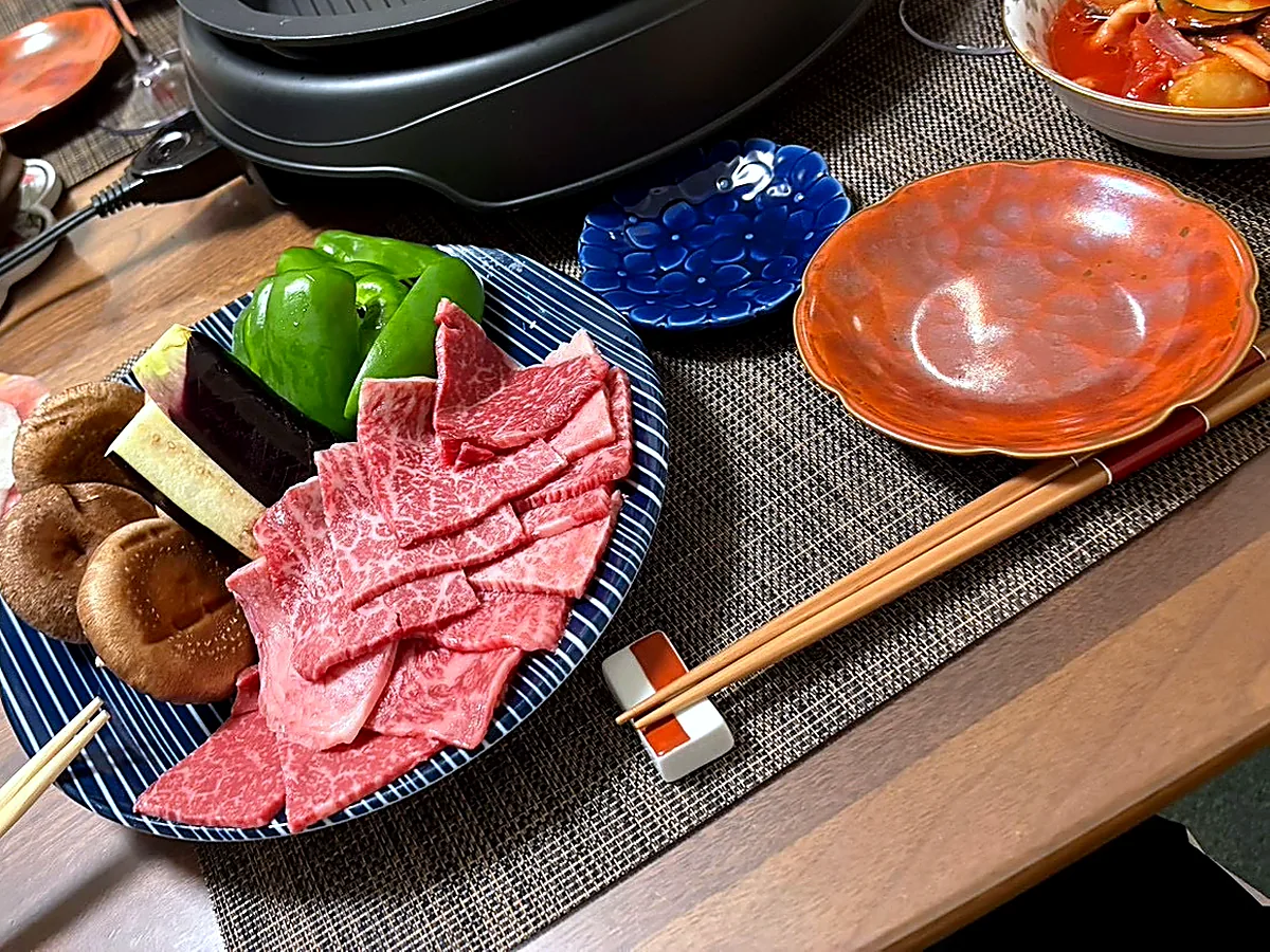 Raw beef and pork slices, shiitake mushrooms, green peppers and eggplant on plates beside a tabletop grill