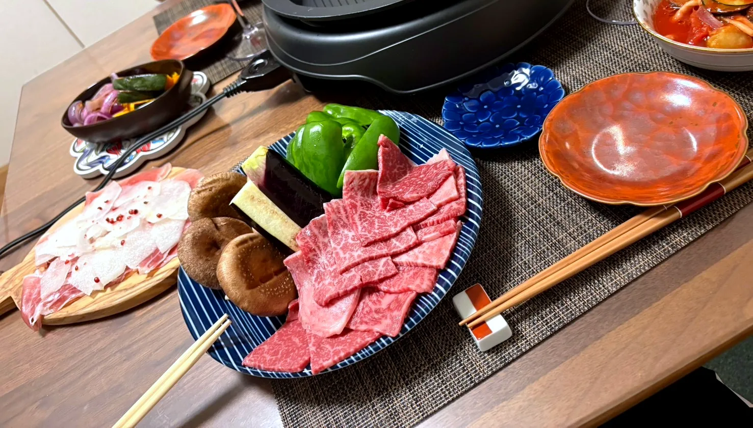 Raw beef and pork slices, shiitake mushrooms, green peppers and eggplant on plates beside a tabletop grill